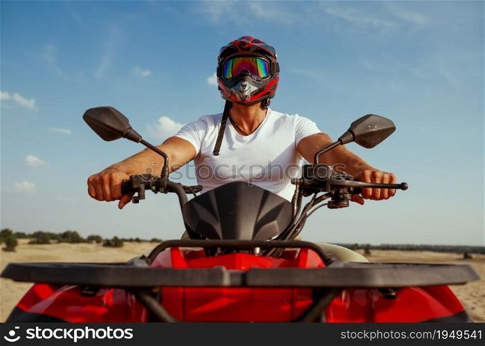 Man in helmet poses on atv in desert, front view. Male person on quad bike, sandy race, dune safari in hot sunny day, 4x4 extreme adventure, quad-biking. Man in helmet poses on atv in desert, front view