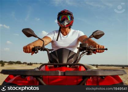 Man in helmet poses on atv in desert, front view. Male person on quad bike, sandy race, dune safari in hot sunny day, 4x4 extreme adventure, quad-biking. Man in helmet poses on atv in desert, front view