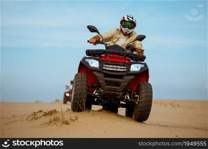 Man in helmet rides on atv, downhill riding in desert sands, action view. Male person on quad bike, sandy race, dune safari in hot sunny day, 4x4 extreme adventure, quad-biking. Man in helmet rides on atv in desert, action view