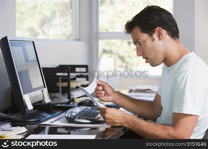 Man in home office using computer holding paperwork and looking shocked
