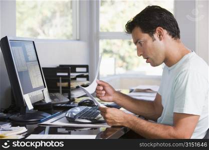 Man in home office using computer holding paperwork and looking shocked