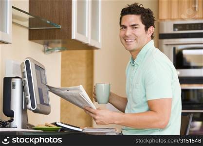 Man in kitchen with computer holding newspaper and coffee smiling