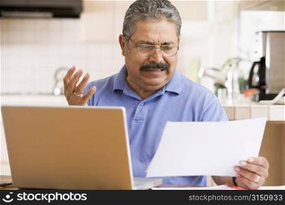 Man in kitchen with laptop and paperwork frustrated
