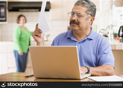 Man in kitchen with laptop and paperwork with woman in background
