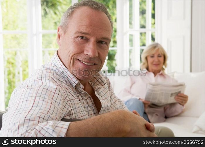 Man in living room smiling with woman in background reading newspaper