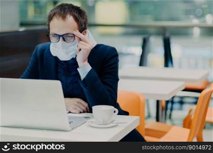 Man in medical mask and spectacles, sits in front of modern laptop computer, poses in cozy cafeteria, reads news about coronavirus, concerned about health, drinks coffee. Epidemic quarantine