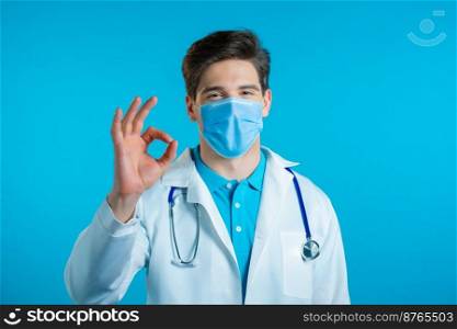 Man in medical white coat and mask showing OK sign over blue background. Positive doctor smiles to camera. Winner. Success. Body language. High quality photo. Man in medical white coat and mask showing OK sign over blue background. Positive doctor smiles to camera. Winner. Success. Body language.
