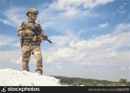 Man in military camouflage uniform and mask, equipped tactical ammunition, standing on sand dune with service rifle replica in hands, cloudy sky on background. Airsoft player taking part in war games. Airsoft player witt gun taking part in war games