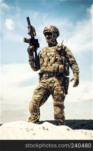 Man in military camouflage uniform and mask, equipped tactical ammunition, standing on sand dune with service rifle replica in hands, cloudy sky on background. Airsoft player taking part in war games. Airsoft player witt gun taking part in war games