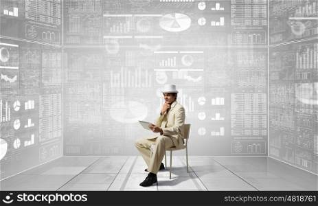 Man in suit and hat use tablet. Young businessman sitting in chair and using tablet