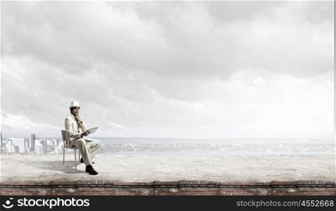 Man in suit and hat use tablet. Young thoughtful businessman sitting in chair on top and using tablet