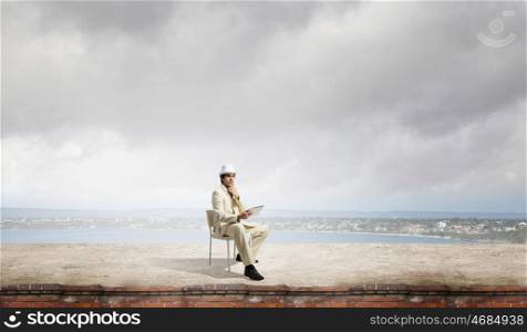 Man in suit and hat use tablet. Young thoughtful businessman sitting in chair on top and using tablet