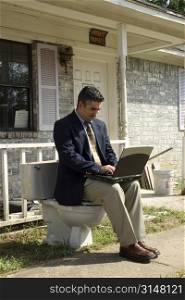 Man in suit with laptop sitting on toliet outside home.
