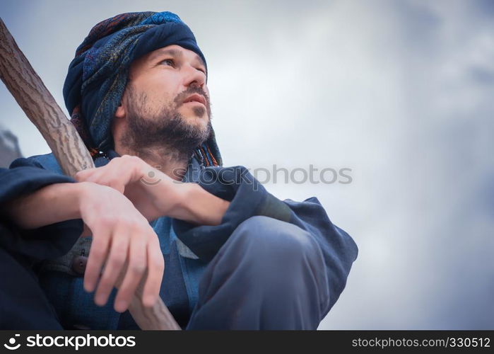 Man in the blue patterned turban with a stick sitting and seriously looking to the side on a light background. Selective focus on face.. Portrait of a Man In Blue Turban