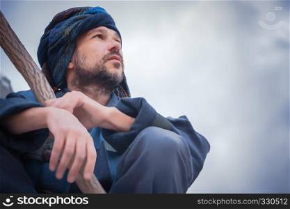 Man in the blue patterned turban with a stick sitting and seriously looking to the side on a light background. Selective focus on face.. Portrait of a Man In Blue Turban