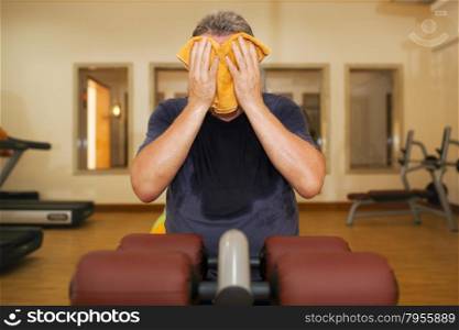 Man in the gymnasium after workout. He is sweaty, exhausted and wiping the face with a towel