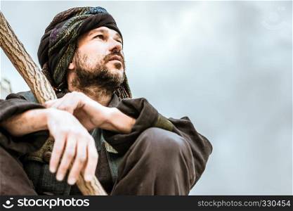 Man in the patterned turban with a stick sitting and seriously looking to the side on a light background. Selective focus on face, toned.. Portrait of a Man In Turban