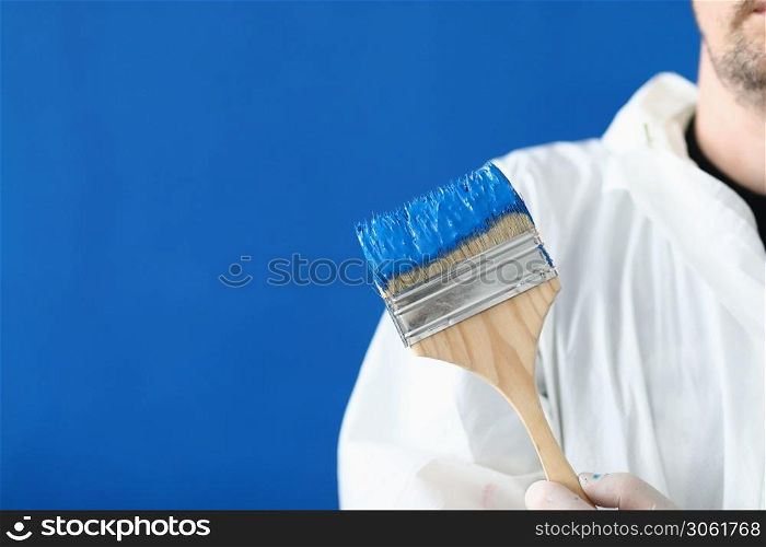 Man in white protective suit hold paintbrush in his hand. Wooden brush smeared with blue paint against blue wall.. Man in white protective suit hold paintbrush in his hand.