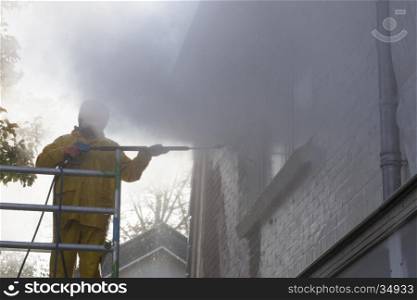 man in yellow rain suit cleans paint from brick wall of house facade with pressure washer while standing on scaffolding