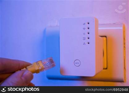 Man insert ethernet cable into WiFi extender device which is in electrical socket on the wall. The device is in access point mode that help to extend wireless network in home or office.