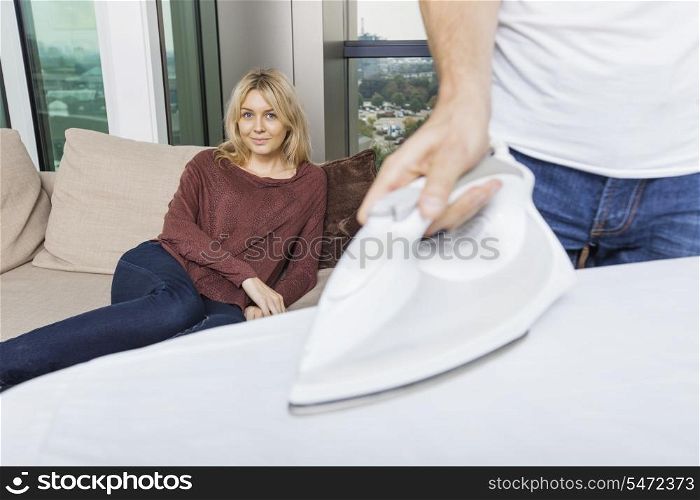 Man ironing shirt while woman relaxing on sofa at home
