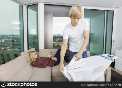 Man ironing shirt while woman relaxing on sofa at home