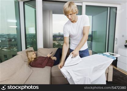 Man ironing shirt while woman relaxing on sofa at home