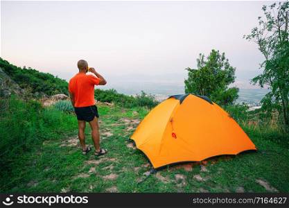 Man is drinking from a cup near an orange tent standing on a hill above the valley. The concept of freedom and travel. Man is drinking from cup near an orange tent standing on hill