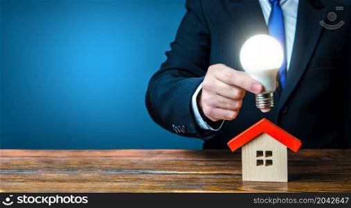 Man is holding a glowing light bulb over the house. Smart home concept. Provision of housing with electricity and communications. Deal contract with supply utilities companies. Pay electricity bills