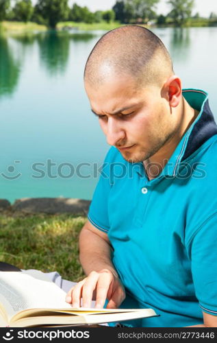 man is sitting outside in a park with lake and reading a book