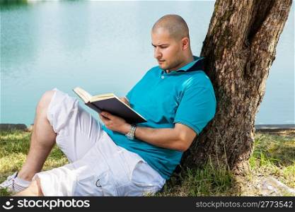 man is sitting outside in a park with lake and reading a book