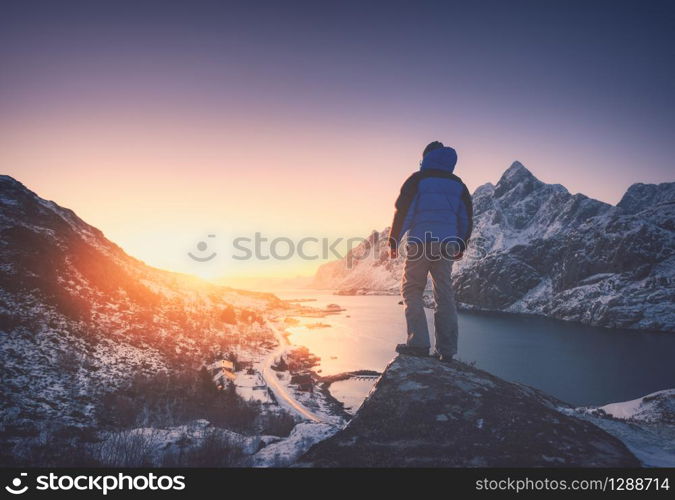 Man is standing on the mountain peak against snowy mountains, fjord at colorful sunset in winter. Young man on the stone, sea coast and rocks, blue sky in Lofoten Islands, Norway. Travel and hiking