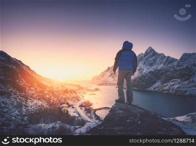 Man is standing on the mountain peak against snowy mountains, fjord at colorful sunset in winter. Young man on the stone, sea coast and rocks, blue sky in Lofoten Islands, Norway. Travel and hiking