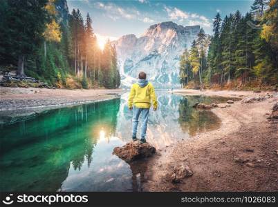 Man is standing on the stone on Braies lake at sunrise in autumn. Dolomites, Italy. Landscape with guy, mountains, beautiful reflection in water, colorful trees, blue sky with sun. Forest in fall. Man is standing on the stone on Braies lake at sunrise in autumn