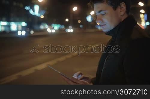 Man is waiting for a bus on a bus stop. He&acute;s holding a tablet in hands and writing letters.