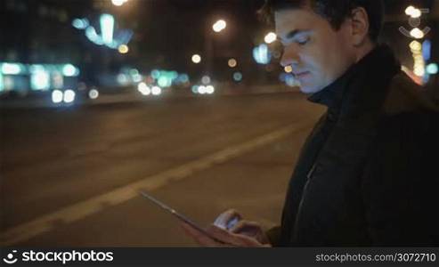 Man is waiting for a bus on a bus stop. He&acute;s holding a tablet in hands and writing letters.