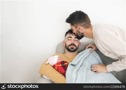 man kissing his boyfriend s forehead sleeping sofa with baby against white wall