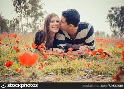 Man kissing woman and she has a toothy smile while they laying on the grass in a field of red poppies.