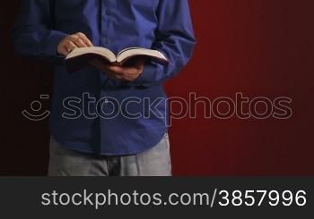 Man leans against a red wall and reads his bible, turning pages then putting his hand in his pocket