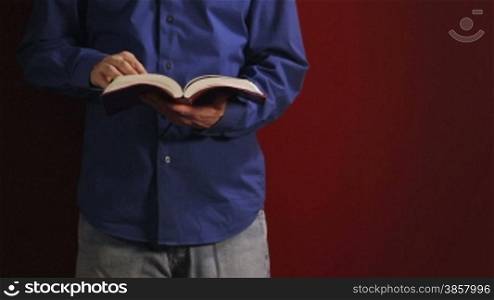 Man leans against a red wall and reads his bible, turning pages then putting his hand in his pocket