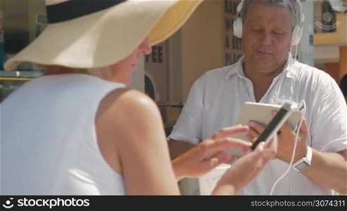 Man listening to music on his tablet and showing something to his wife who using smartphone. Summertime.