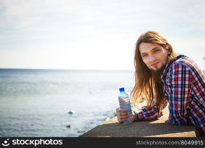 Man long hair relaxing by seaside drinking water. Man long hair wearing plaid shirt relaxing by seaside drinking water at summer sunny day