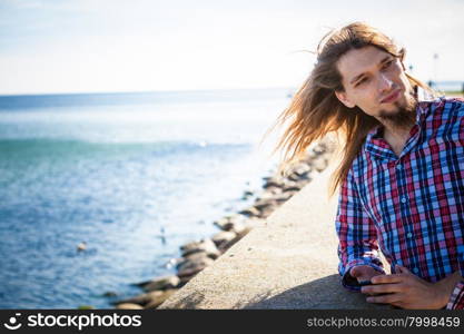 Man long hair relaxing by seaside . Man bearded long hair wearing plaid shirt casual style relaxing by seaside at summer sunny windy day