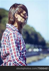 Man long hair relaxing by seaside . Man bearded long hair wearing plaid shirt casual style relaxing by seaside at summer sunny windy day