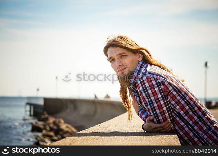 Man long hair relaxing by seaside . Man bearded long hair wearing plaid shirt casual style relaxing by seaside at summer sunny windy day