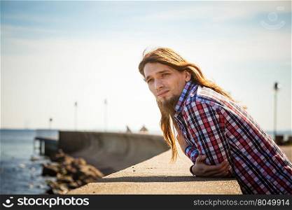 Man long hair relaxing by seaside . Man bearded long hair wearing plaid shirt casual style relaxing by seaside at summer sunny windy day