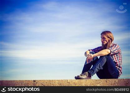 Man long hair relaxing outdoor sky background. Man long hair wearing plaid shirt relaxing outdoor sitting on concrete wall at sunny windy day against blue sky