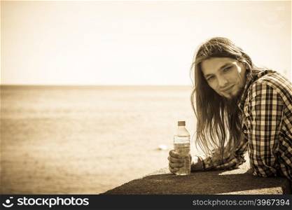 Man long hair wearing plaid shirt relaxing by seaside drinking water at summer sunny day. Man long hair relaxing by seaside drinking water
