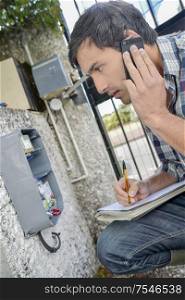 Man looking at exterior electrical box, on telephone taking notes