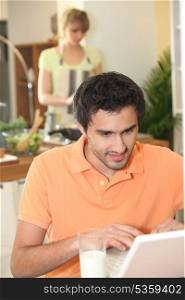 Man looking at his laptop while his wife prepares dinner